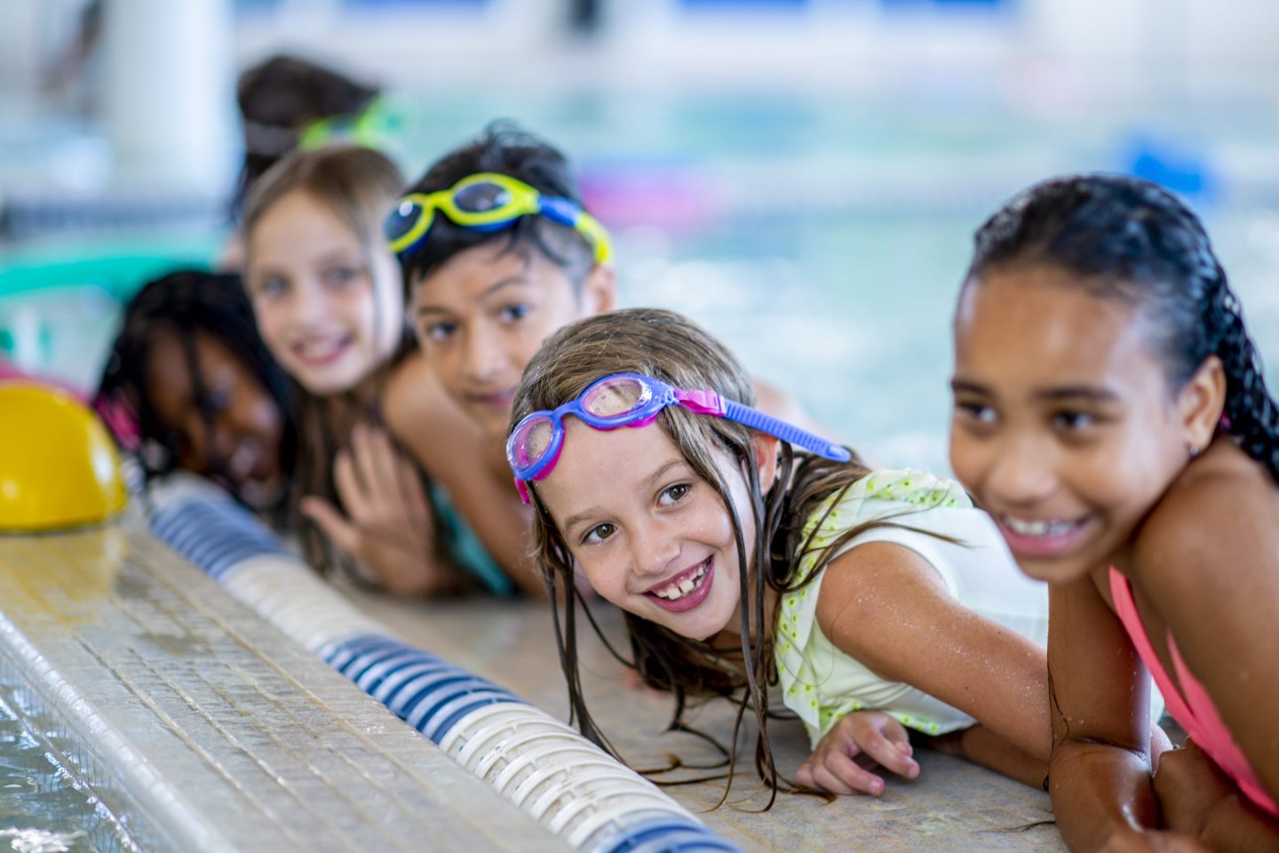 A multi-ethnic group of kids are indoors in a pool. Some of them are wearing goggles and smiling at the camera.